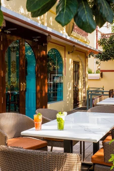Agan Cafe outdoor seating area featuring elegant wicker chairs, white marble tables, colourful refreshing drinks, and a charming yellow exterior with turquoise arched windows, surrounded by lush green foliage in a warm, inviting ambience.
