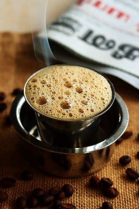 Traditional South Indian filter coffee served hot in a stainless steel tumbler and davara, topped with frothy golden-brown foam, with coffee beans scattered on a rustic burlap surface and a newspaper in the background.