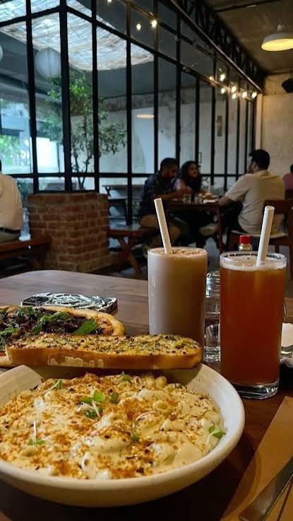 A cosy cafe interior with floor-to-ceiling glass windows, warm hanging lights, and wooden tables, featuring creamy pasta, garlic bread, pizza, and refreshing iced beverages served in a relaxed dining atmosphere.
